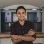 Positive Indian in casual outfit standing with crossed arms near glass fence while smiling and looking at camera in modern corridor